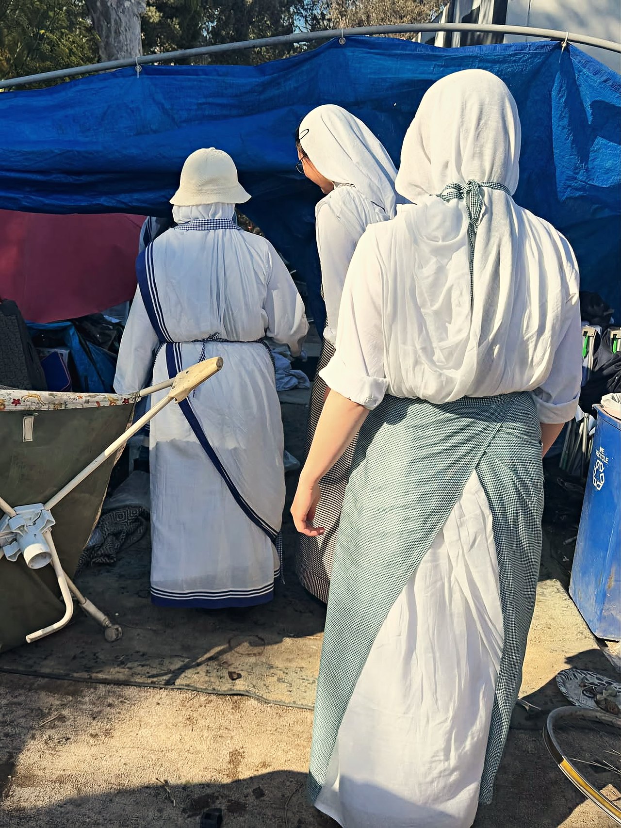 Missionaries of Charity sisters in white and blue habits at a tarp-covered encampment.