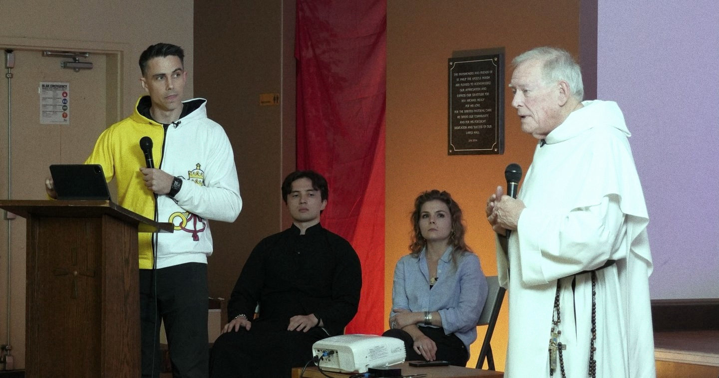 Fr. Michael Sweeney, OP speaking into a handheld microphone in his white Dominican habit while Sean Bryan stands at the lectern, a deep red curtain behind them.